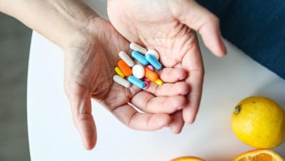 Manos sosteniendo varios suplementos y vitaminas de colores junto a una naranja
