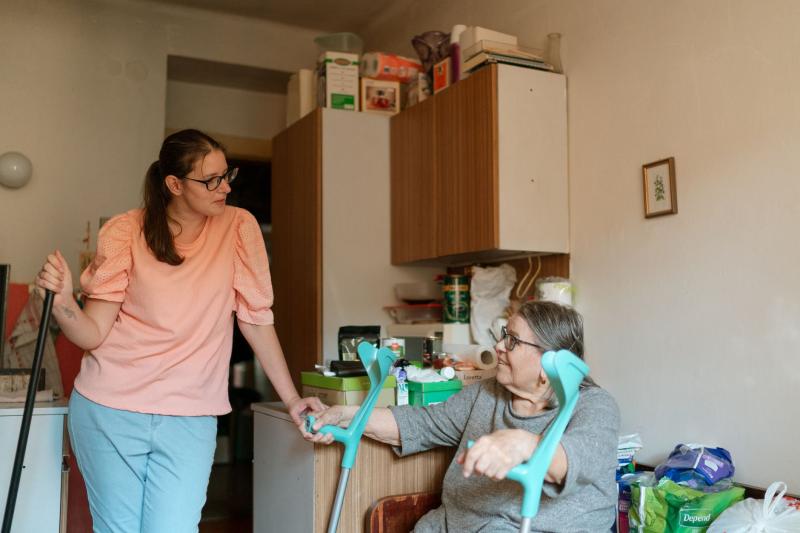 Mujer mayor con muletas apoyada en la encimera de su cocina, atendida por una mujer que la ayuda en casa
